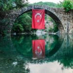 A serene view of a stone bridge with a Turkish flag reflecting on the water below.